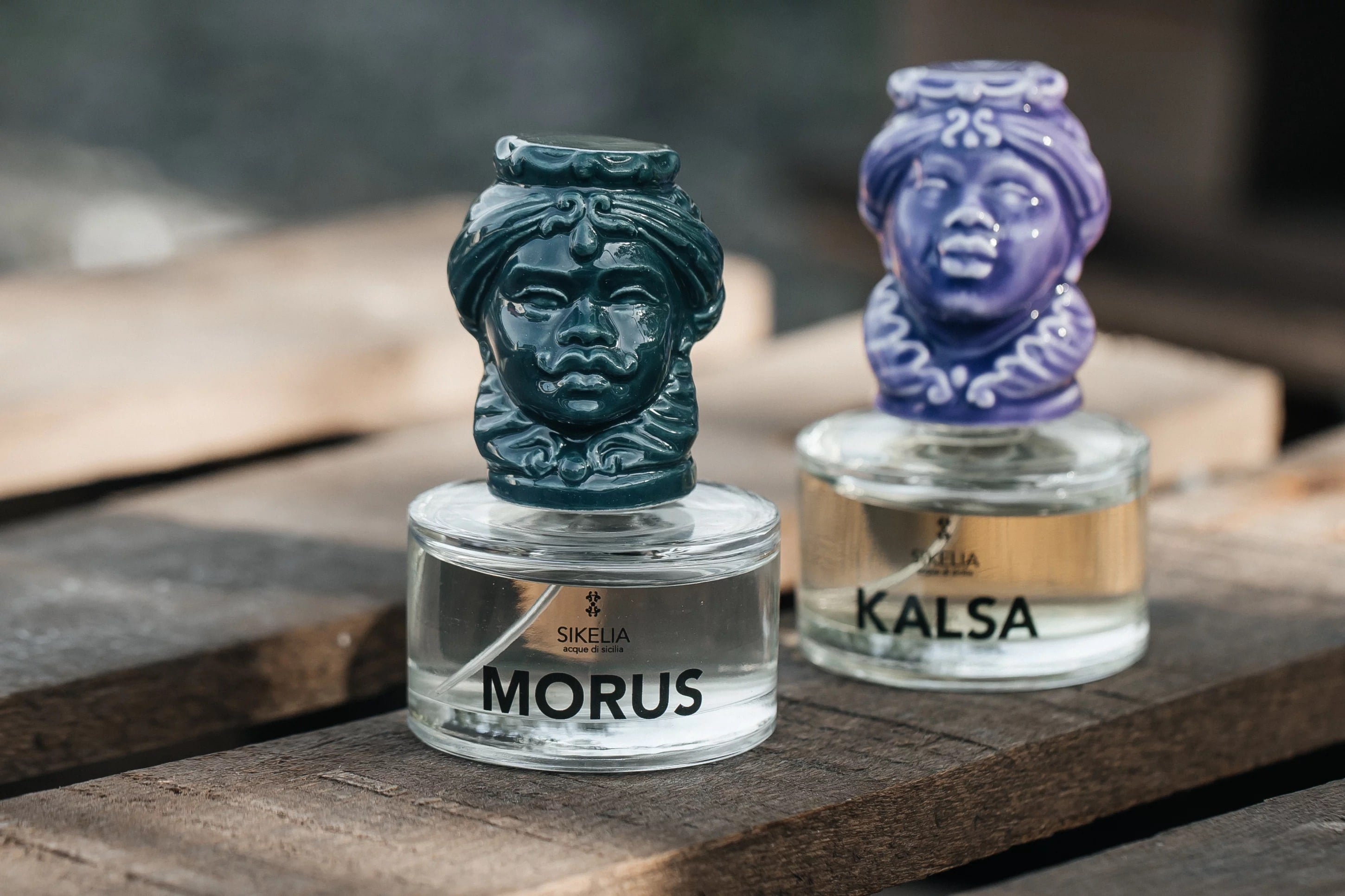 Two Sikelia perfume bottles labeled "MORUS" and "KALSA," each with a sculpted decorative stopper resembling a human face, placed on a wooden surface.